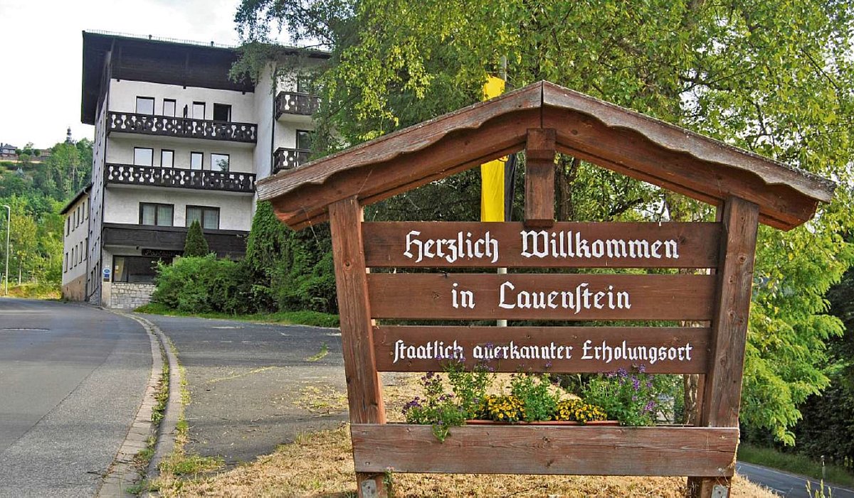 Im Vordergrund ein großes Holzschild mit dem Text "Herzlich Willkommen in Lauenstein". Im Hintergrund ist ein großes, weißes Gebäude mit dunkelbrauner Holzverkleidung zu sehen.