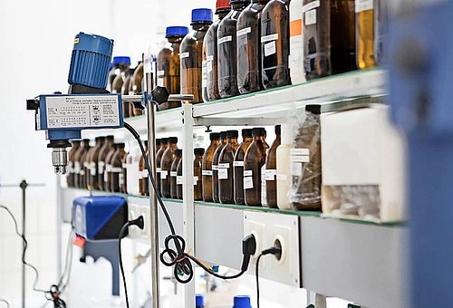 An einem technischen Arbeitsplatz stehen zahlreiche brauen Flaschen mit verschiedenen Etiketten auf einem Regal nebeneinander.