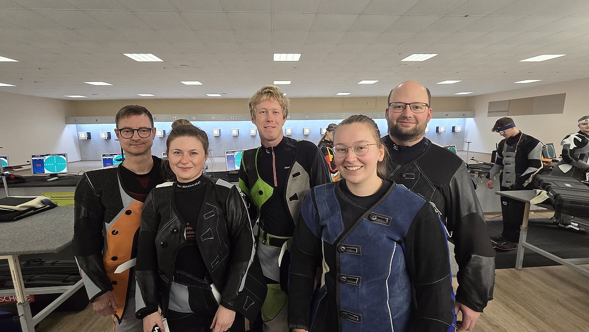 5 Menschen in Sportschützenkleidung in einem Schützenraum. Im Hintergrund sind Schießstände zu sehen.