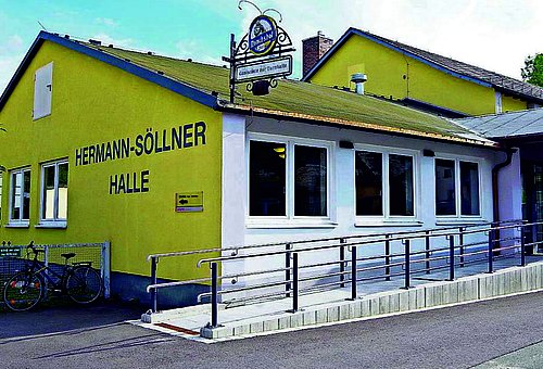 Zu sehen ist die neu gestaltete Hermann-Söllner-Halle. Die Front strahlt in einem hellen Gelb mit Schriftzug Hermann-Söllner-Halle. Rechts davon ist der barrierefreie Zugang zur Halle.