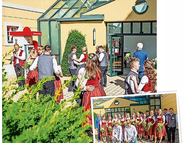 Tanzpaare in Trachtenkleidung tanzen vor einem gelben Gebäude mit Glasflächen. 