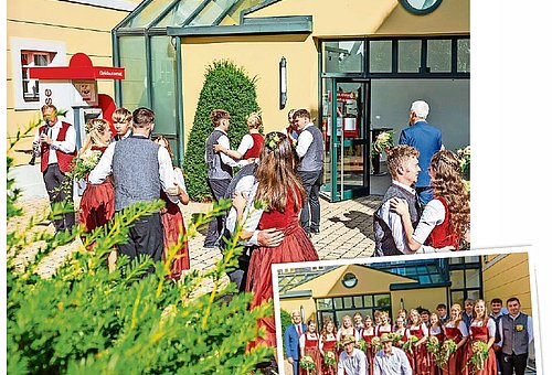Tanzpaare in Trachtenkleidung tanzen vor einem gelben Gebäude mit Glasflächen. 