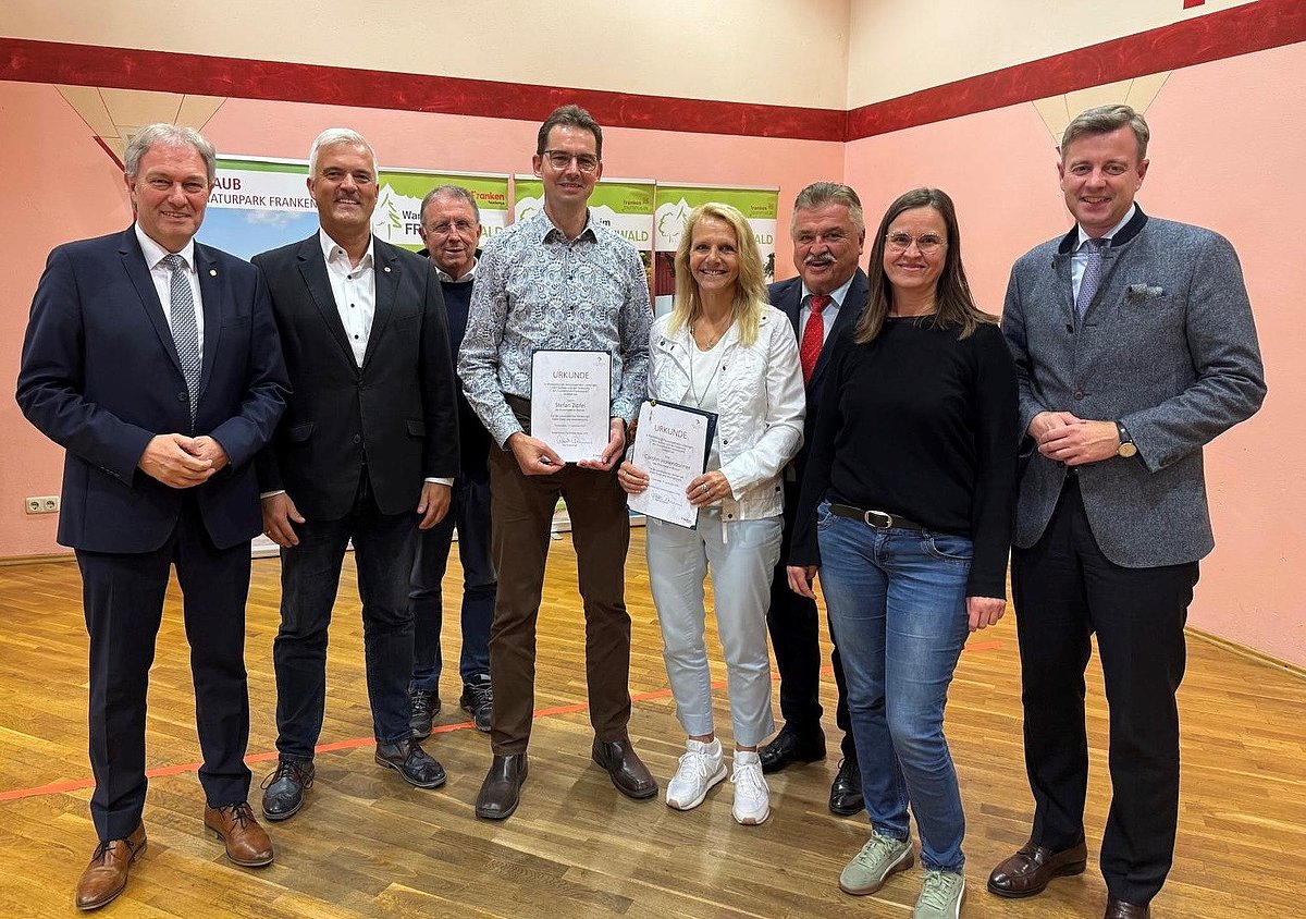 Eine Gruppe von Menschen, darunter zwei Frauen stehen in einem Halbkreis und lächeln in die Kamera. Ein Mann und eine Frau, die in der Mitte stehen halten jeweils eine Urkunde in der Hand.