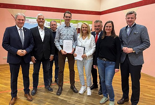 Eine Gruppe von Menschen, darunter zwei Frauen stehen in einem Halbkreis und lächeln in die Kamera. Ein Mann und eine Frau, die in der Mitte stehen halten jeweils eine Urkunde in der Hand.
