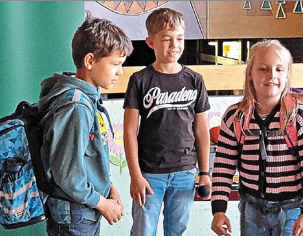 Drei Schulkinder davon ein Mädchen stehen zusammen. Das Mädchen und ein Junge haben Schulränze auf dem Rücken. Sie stehen scheinbar in einem Klassenzimmer. 