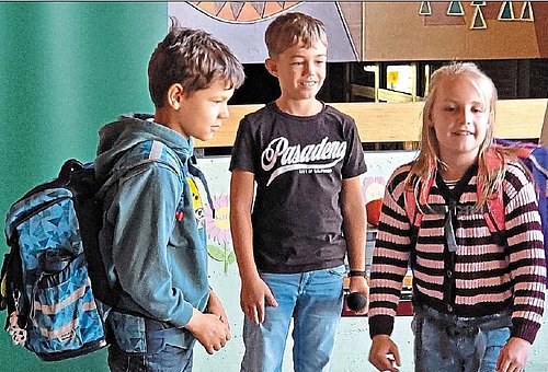 Drei Schulkinder davon ein Mädchen stehen zusammen. Das Mädchen und ein Junge haben Schulränze auf dem Rücken. Sie stehen scheinbar in einem Klassenzimmer. 