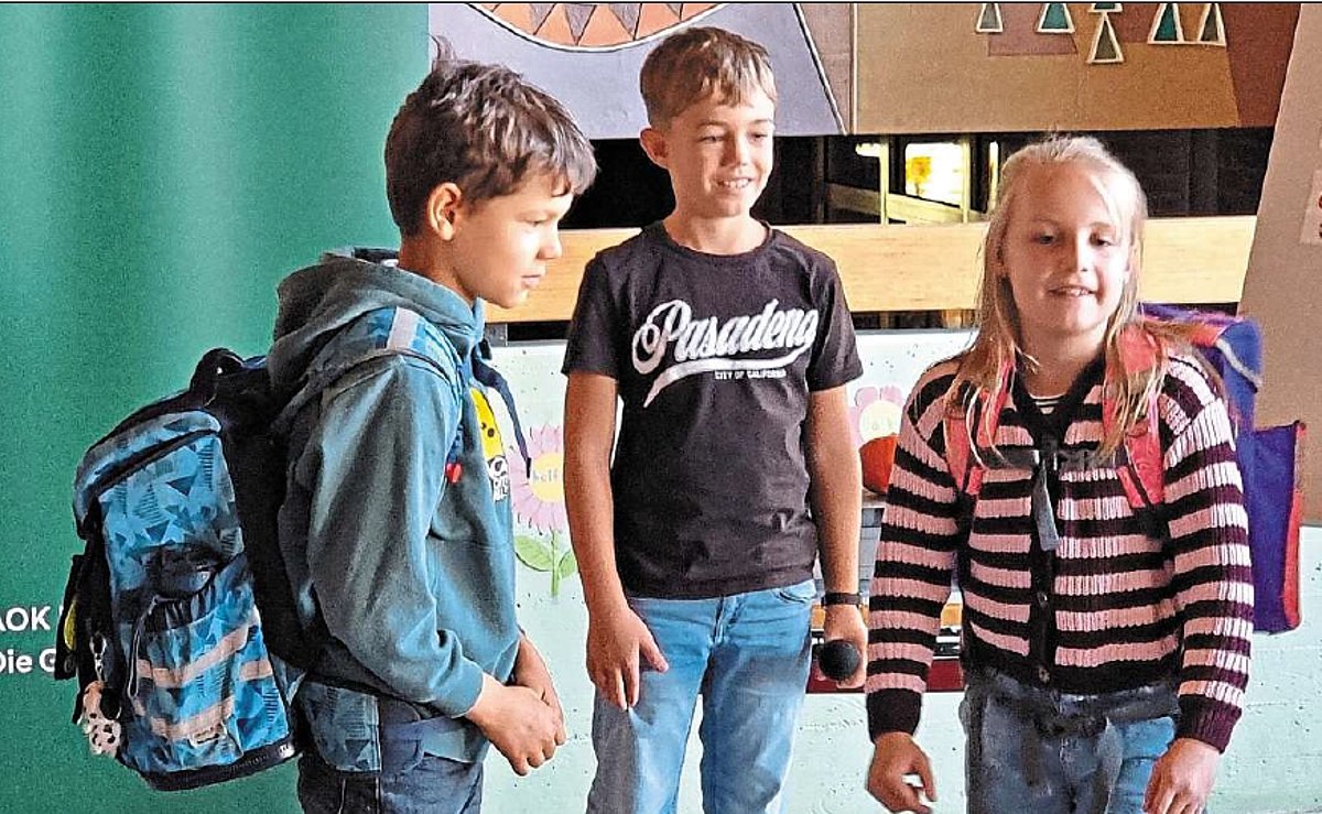 Drei Schulkinder davon ein Mädchen stehen zusammen. Das Mädchen und ein Junge haben Schulränze auf dem Rücken. Sie stehen scheinbar in einem Klassenzimmer.