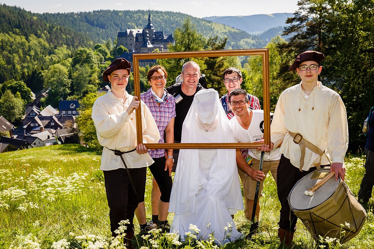 Das Foto zeigt zwei junge Männer, die einen überdimensional großen leeren Bilderrahmen halten. Einige Personen und die weiße Frau schauen durch diesen Fotorahmen zum Fotografen.