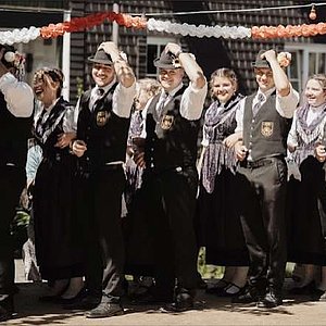 Eine Gruppe von jungen Leuten,gekleidet mit historischer Tracht, stehen  hintereinander in einer Reihe als Paar. Im Hintergrund sind noch bunte Girlanden zu sehen.