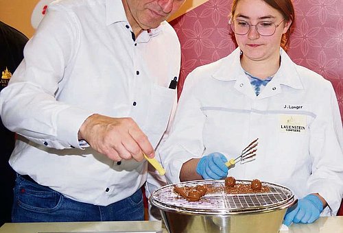 Das Fot zeigt Landrat Klaus Löffler beim Garnieren einer runden Praline auf einem Gitterrost. Eine junge Frau im weißen Kittel und mit Handschuhen steht daneben und schaut ihm zu.