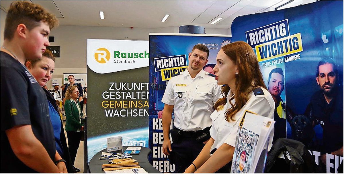 Eine Polizistin steht vor einer Infowand und ist in einem Gespräch mit einem jungen Mann. Im Hintergrund ist ein weiterer Polizist in Uniform zu sehen, der zu ihr schaut und lächelt. 