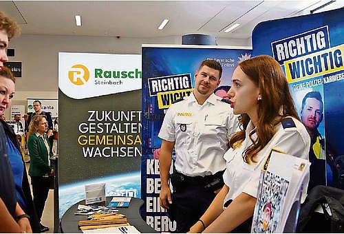 Eine Polizistin steht vor einer Infowand und ist in einem Gespräch mit einem jungen Mann. Im Hintergrund ist ein weiterer Polizist in Uniform zu sehen, der zu ihr schaut und lächelt. 