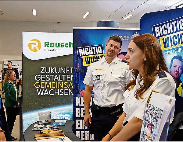 Eine Polizistin steht vor einer Infowand und ist in einem Gespräch mit einem jungen Mann. Im Hintergrund ist ein weiterer Polizist in Uniform zu sehen, der zu ihr schaut und lächelt.
