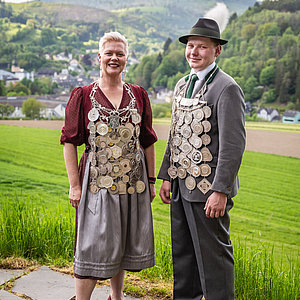 Links ist die amtierende Schützenkönigin in einem dunkelrotem Dirndl mit der Kette zu sehen. Rechts neben ihr steht der Jungschützenkönig im traditionellen Schützenanzug und Hut. Auch er trägt stolz seine Königskette.