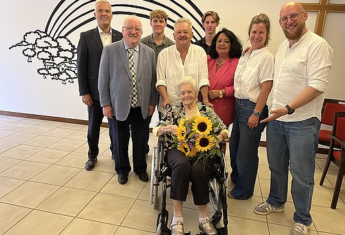 Eine Seniorin sitzt in einem Rollstuhl, hat einen Sonnenblumenstrauß und lächelt in die Kamera. Hinter ihr stehen Männer und Frauen - ihre Familie sowie Bürgermeister Timo Ehrhardt und Landrat-Stellvertreter Bernd Steger.
