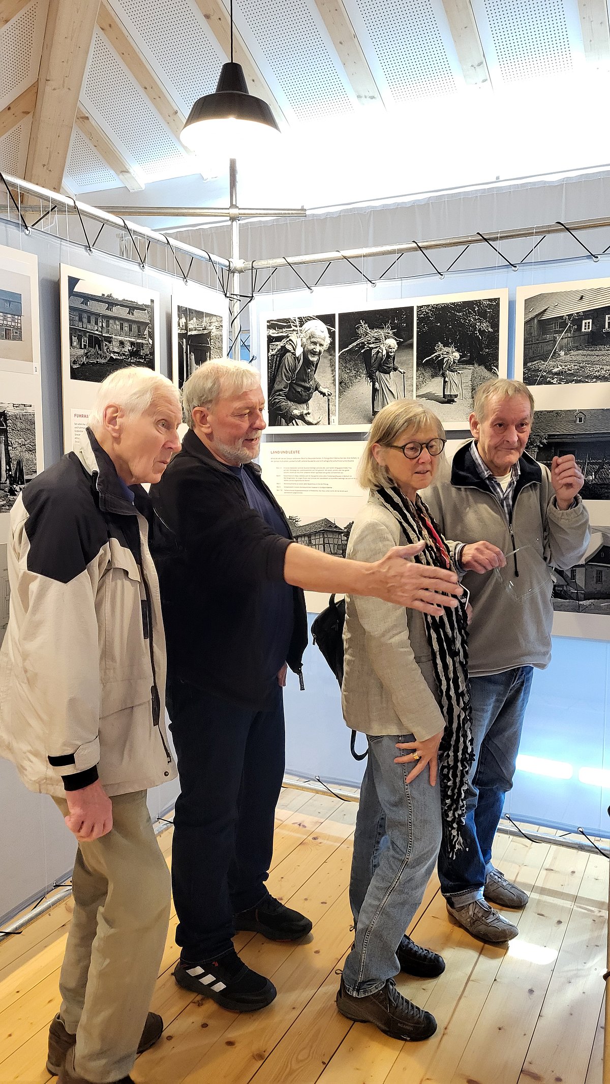 Drei Männer und eine Frau stehen an Fotowänden in einem Gebäude. Einer der Männer deutet mit seiner rechten Hand auf eine Foto. Die anderen schauen interessiert dorthin.