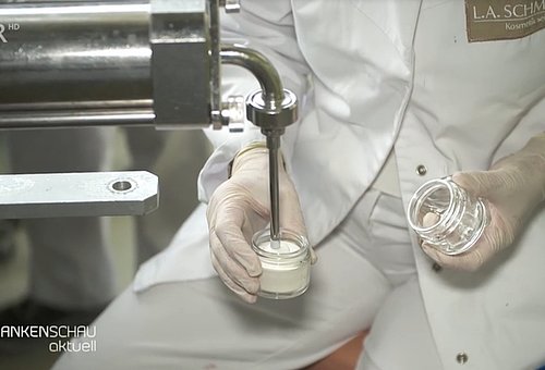 Eine Frau in weißer Arbeitskleidung mit Handschuhen befüllt an einer Maschine Cremedöschen.