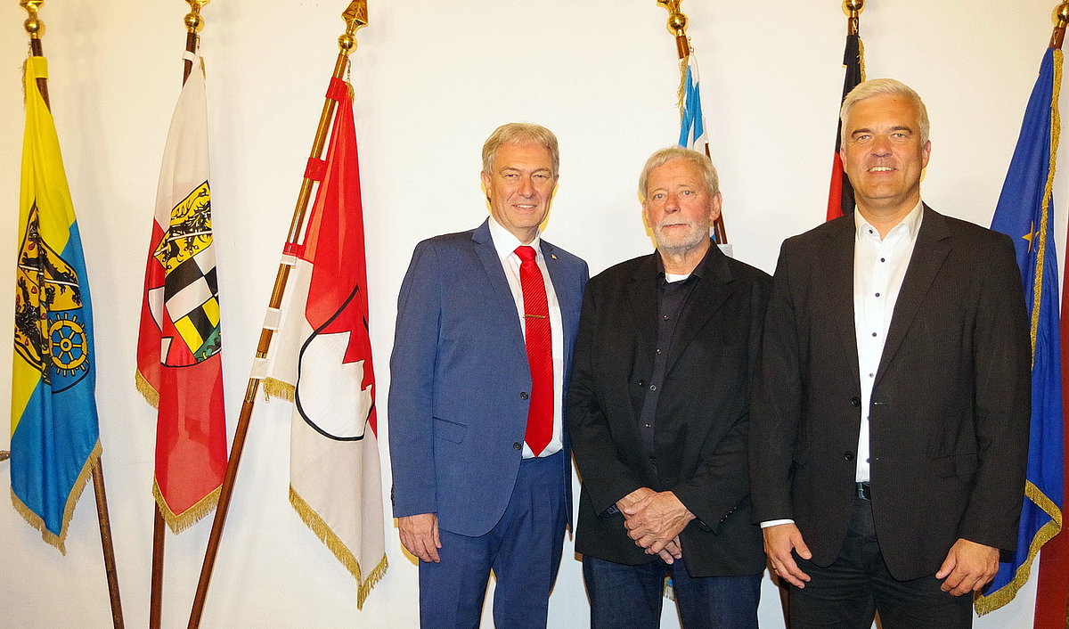 Vor einer Reihe von verschiedenen Fahnen stehen drei Männer nebeneinander. Zwei davon in schwarzen Anzügen, der dritte Mann trägt einen blauen Anzug und eine rote Krawatte.