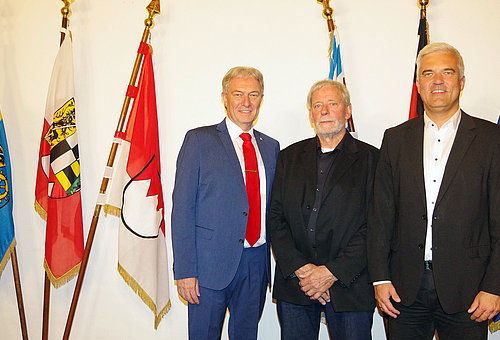 Vor einer Reihe von verschiedenen Fahnen stehen drei Männer nebeneinander. Zwei davon in schwarzen Anzügen, der dritte Mann trägt einen blauen Anzug und eine rote Krawatte. 