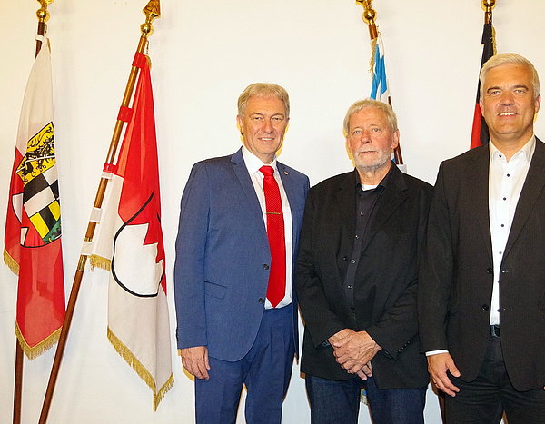 Vor einer Reihe von verschiedenen Fahnen stehen drei Männer nebeneinander. Zwei davon in schwarzen Anzügen, der dritte Mann trägt einen blauen Anzug und eine rote Krawatte.