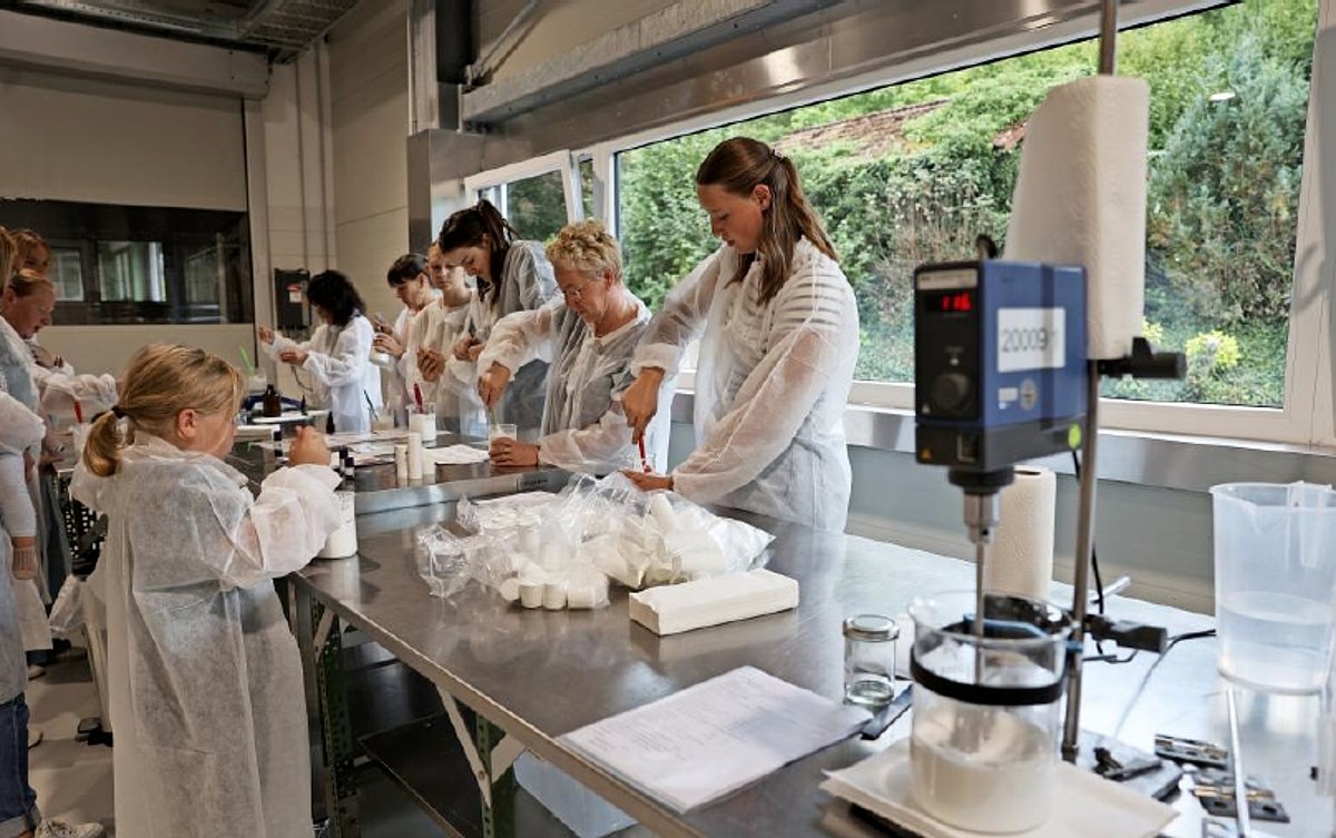 An einem Labortisch mit Laborgeräten stehen zahlreichen Frauen und Mädchen und rühren konzentriert mit Spateln in Laborgläsern. Alle tragen weiße Schutzanzüge.