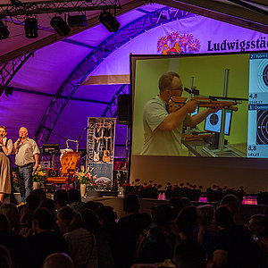 Eine Frau und ein Mann moderieren das Jedermannschießen live im Festzelt. Im Hintergrund sieht man auf einer großen Leinwand einen Schützen, der gerade seinen Königsschuss abgibt.