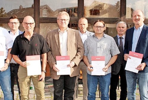 Eine Gruppe von Menschen schauen in die Kamera. Sie stehen in zwei Reihen hintereinander. Die Personen in der vorderen Reihe halten je eine Urkunde in der Hand. Sie alle lächeln.