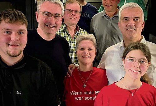 Auf einer Treppe in einem Gebäude steht eine kleine Gruppe von Menschen. Darunter sieben Männer und zwei Frauen. Alle tragen Freizeitkleidung und lächeln in die Kamera.