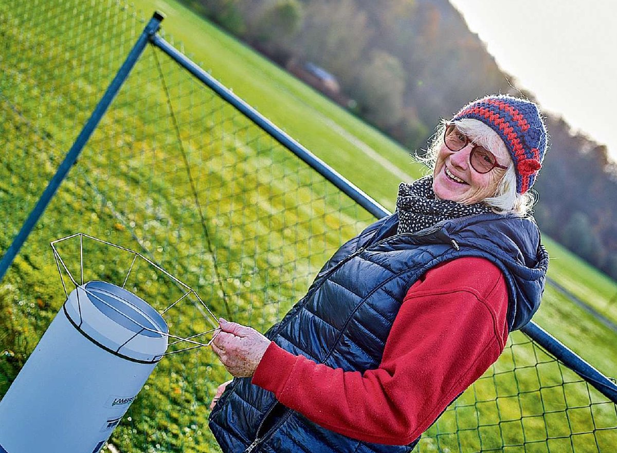 Das Bild zeigt eine Frau mit Pullover, blauer Steppweste und Mütze, die draußen an einer Wetterstation steht. Sie lacht in die Kamera.