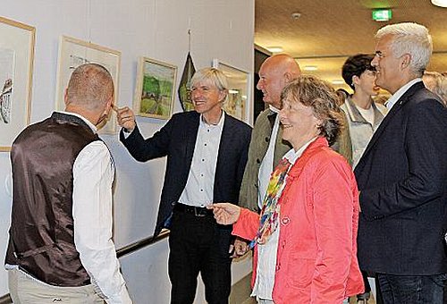 5 Personen, darunter eine Frau in einer roten Jacke, stehen vor einer Wand mit Bildern. Einer der Männer zeigt auf ein Bild - alle Anderen schauen genau auf dieses Bild.