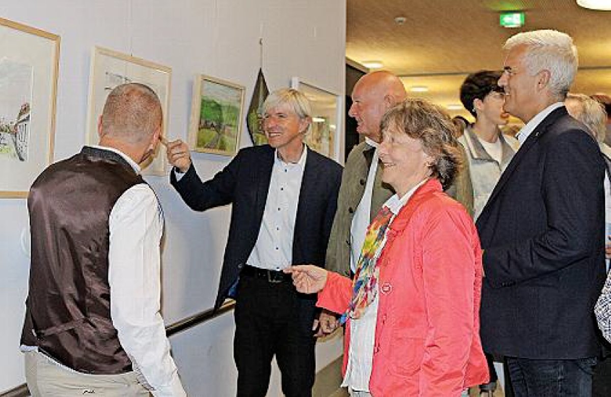 5 Personen, darunter eine Frau in einer roten Jacke, stehen vor einer Wand mit Bildern. Einer der Männer zeigt auf ein Bild - alle Anderen schauen genau auf dieses Bild.