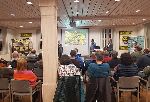 Viele Personen sitzen auf Stühlen in einem große Raum und schauen nach vorn. Sie sind alle von hinten zu sehen. Vorn läuft eine Präsentation und drei Männer stehen dort und schauen ins Publikum.