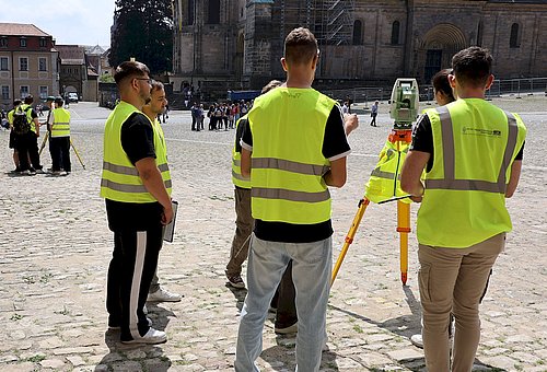Mehrere junge Menschen mit neongrünen Warnwesten stehen auf einem Platz. Zwischen ihnen steht ein Instrument auf einem Dreibein. Es sieht aus wie ein Fernglas oder eine Kamera.