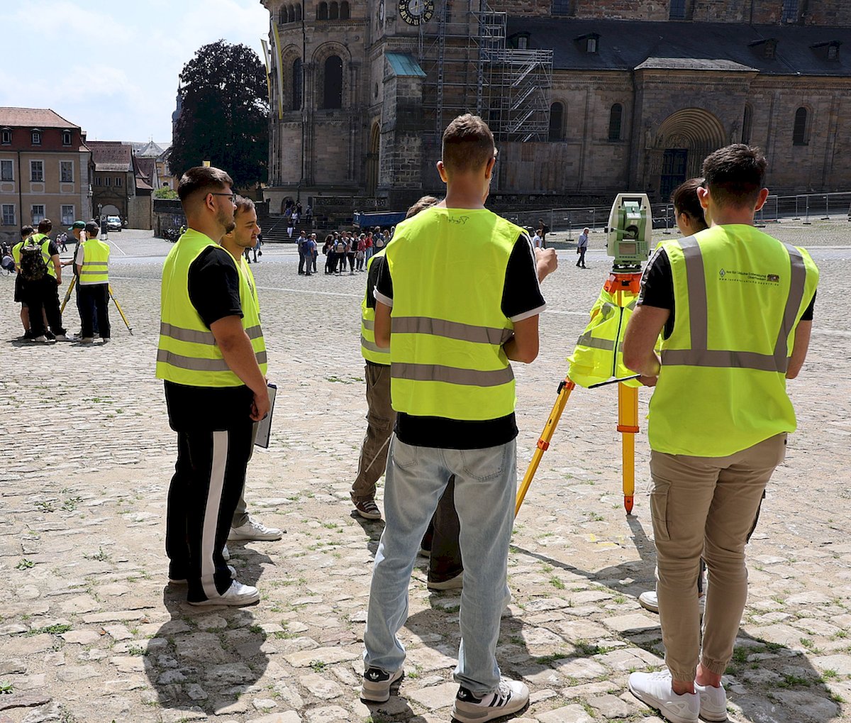 Mehrere junge Menschen mit neongrünen Warnwesten stehen auf einem Platz. Zwischen ihnen steht ein Instrument auf einem Dreibein. Es sieht aus wie ein Fernglas oder eine Kamera.