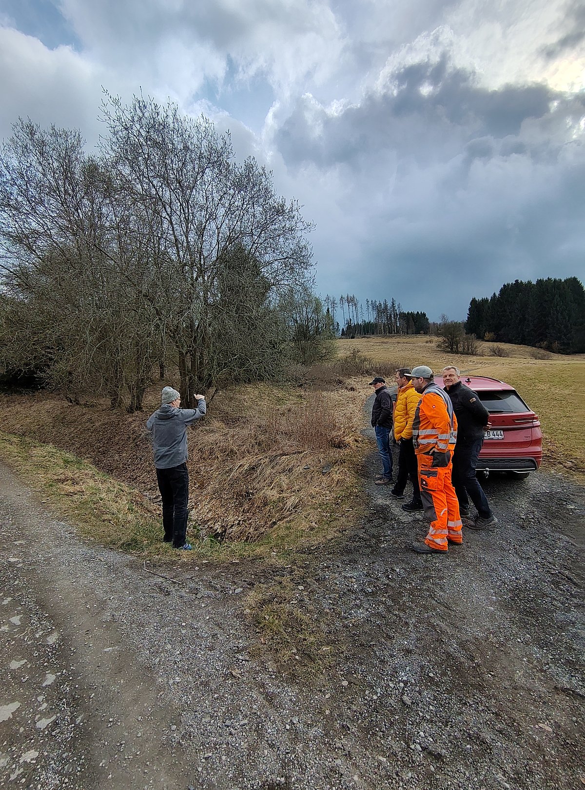 An einem Wiesenweg stehen mehrere Männer und schauen zu einem Mann, der gegenüber der Gruppe steht und scheinbar etwas erklärt.