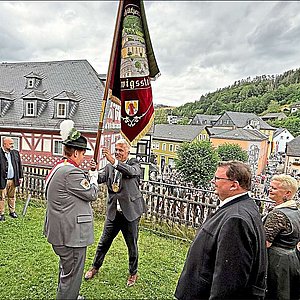 Vor der evang. Kirche mit Blick auf den Marktplatz übergibt Bürgermeister Timo Ehrhardt an ein Mitglied der Schützengesellschaft die Traditionsfahne. Umrahmt werden die beiden von weiteren Ehrengästen links und rechts.