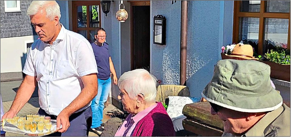 Vor einem Gebäude sind mehrere Personen zu sehen. Ein Mann in einem weißen Hemd serviert ein Tablet mit kleinen Schnapsgläsern, die gefüllt sind.