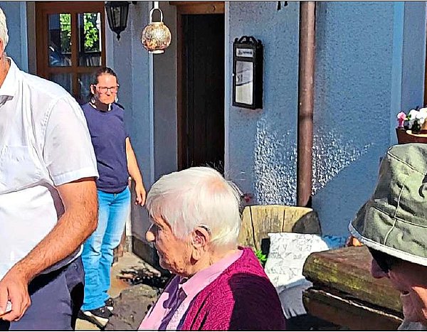 Vor einem Gebäude sind mehrere Personen zu sehen. Ein Mann in einem weißen Hemd serviert ein Tablet mit kleinen Schnapsgläsern, die gefüllt sind.