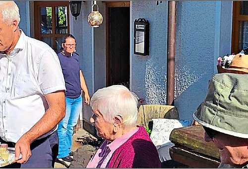 Vor einem Gebäude sind mehrere Personen zu sehen. Ein Mann in einem weißen Hemd serviert ein Tablet mit kleinen Schnapsgläsern, die gefüllt sind. 