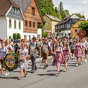 Das Foto zeigt den Schützenumzug. Hier sieht man den Anfang des Zuges. Vorn wird die Schützenscheibe von zwei Kindern getragen. Dahinter laufen Schützenkönig, Jungkönig und Jedermannkönig umrahmt von den Schützenliesln in neuer Tracht. Diese ist dunkelrosa.