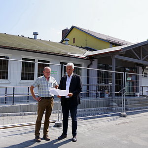 Zwei Männer stehen mit einem Plan in der Hand vor einer Halle, die mit Bauzäunen umstellt ist.