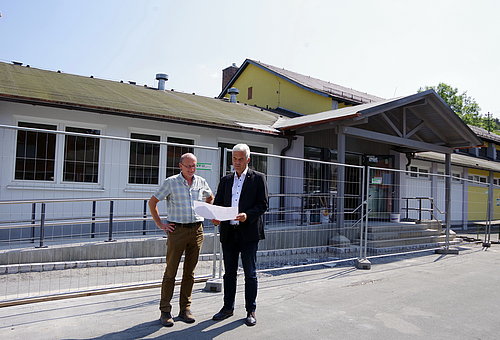 Zwei Männer stehen mit einem Plan in der Hand vor einer Halle, die mit Bauzäunen umstellt ist. 