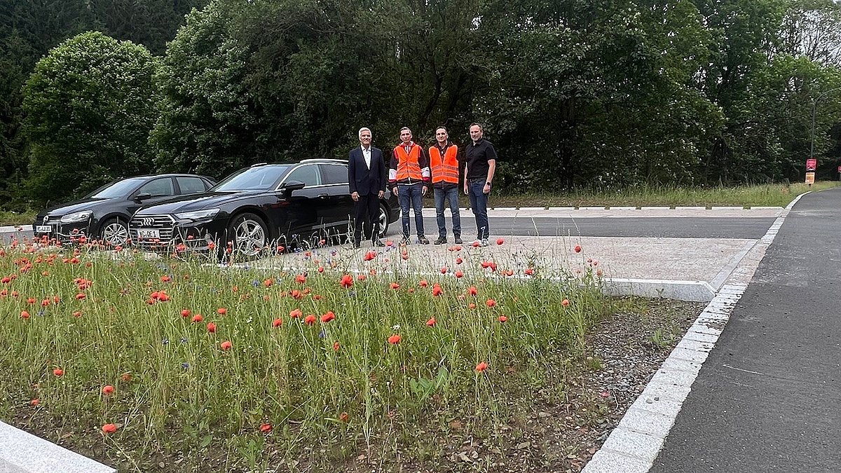 Auf einer neuen Parkplatzfläche mit Grünflächen am Rand stehen zwei parkende Autos und der Bürgermeister sowie drei weitere Männer stehen zusammen.