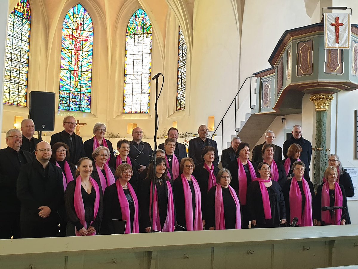 Ein gemischter Chor in einer Kirche. Alle tragen schwarze Kleidung, die Frauen zusätzlich ein pinkfarbenes Halstuch.