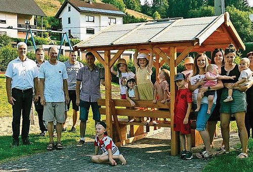 Eine Gruppe von Menschen, darunter viele Kinder, stehen an einer Sitzgarnitur mit Tisch und Bänken, die aus Holz sind und ein Dach haben. Sie alle lächeln in die Kamera.