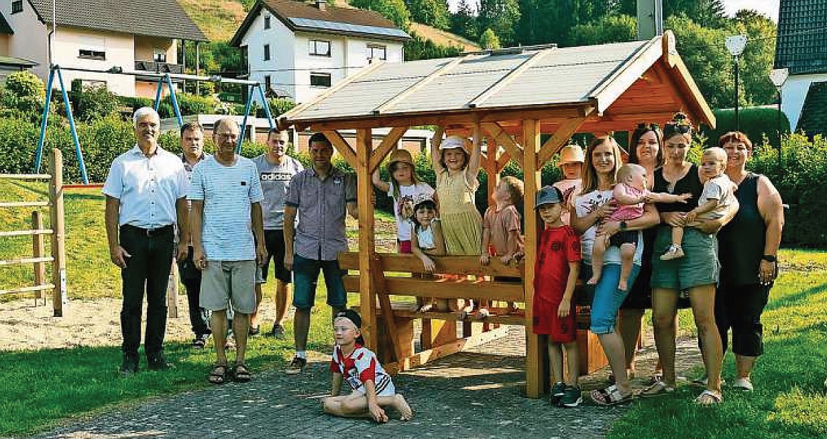 Eine Gruppe von Menschen, darunter viele Kinder, stehen an einer Sitzgarnitur mit Tisch und Bänken, die aus Holz sind und ein Dach haben. Sie alle lächeln in die Kamera.