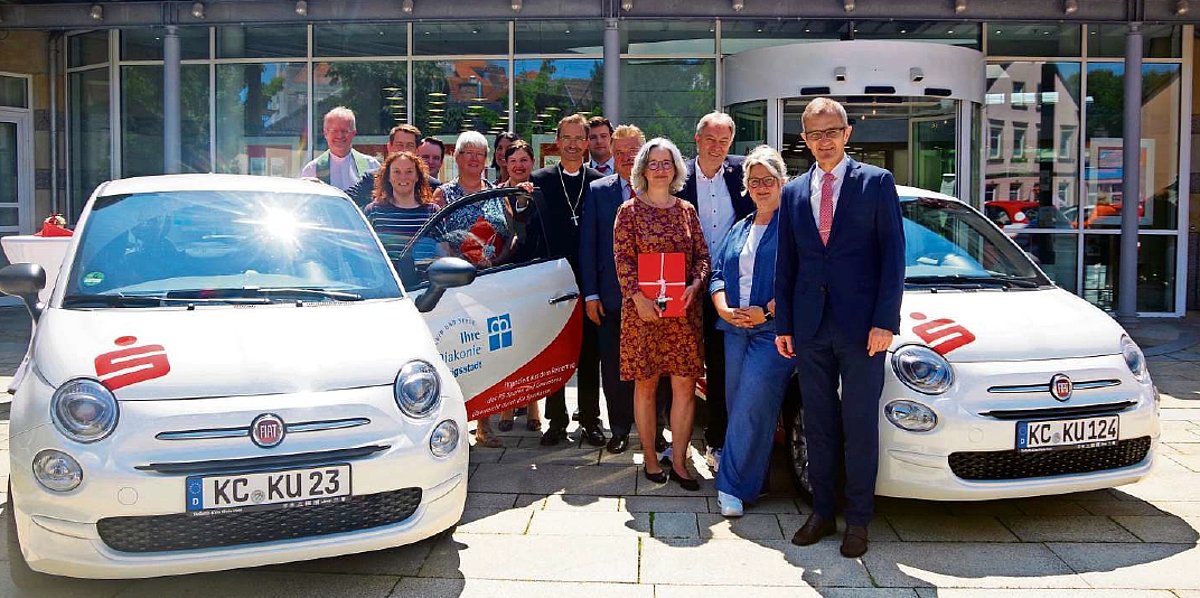 Zwischen den zwei weißen, neuen Kleinwagen steht eine Gruppe von Menschen. Sie alle schauen in die Kamera.