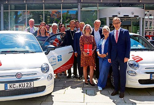 Zwischen den zwei weißen, neuen Kleinwagen steht eine Gruppe von Menschen. Sie alle schauen in die Kamera.