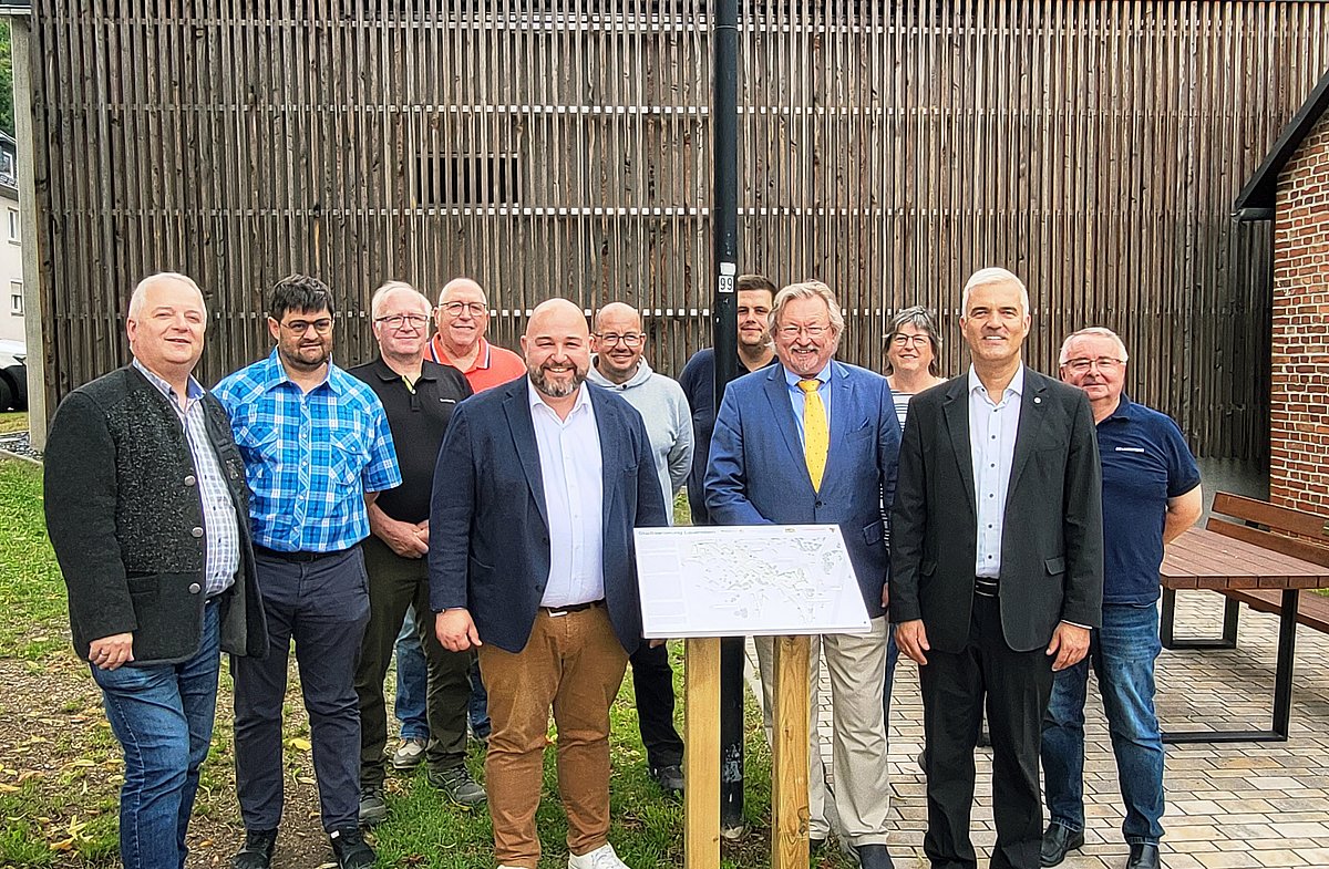 Eine Gruppe von Männern und einer Frau stehen draußen. Sie alle schauen in die Kamera. Vor ihnen steht eine weiße Tafel mit Text und Bildern auf zwei Holzpfosten.