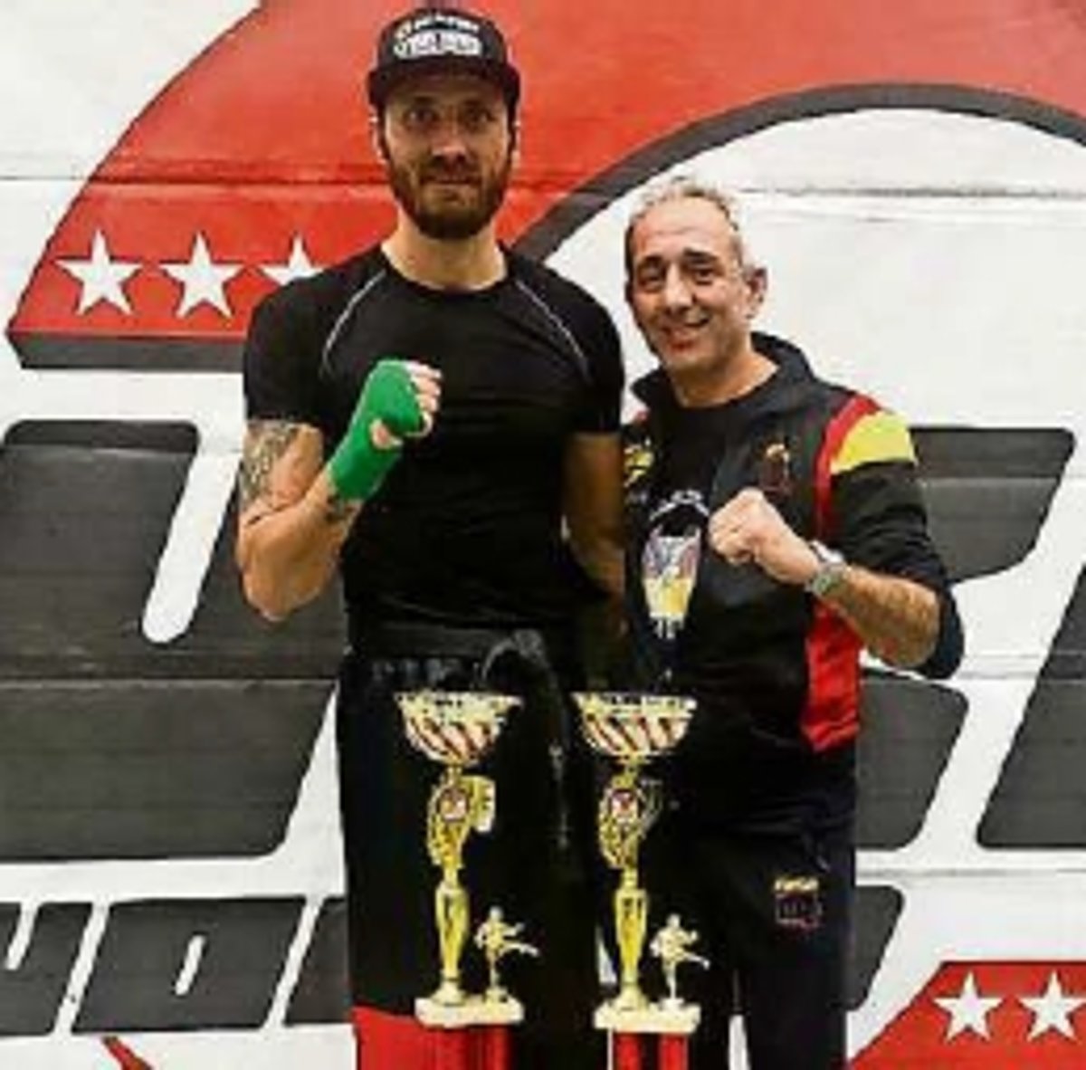 Zwei Männer in Trainingskleidung stehen vor einer rot weißen Fahne, schauen in Kamera und haben jeweils eine Hand zur Faust geballt. Vor ihnen stehen zwei goldene Pokale.